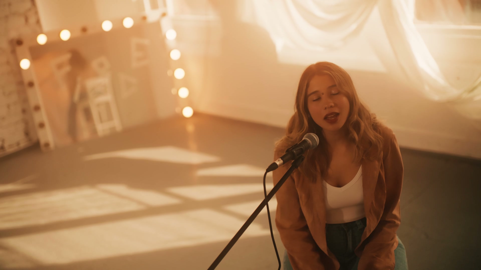 Lilliane singing at a microphone, with warm window light and shadows across the studio floor.