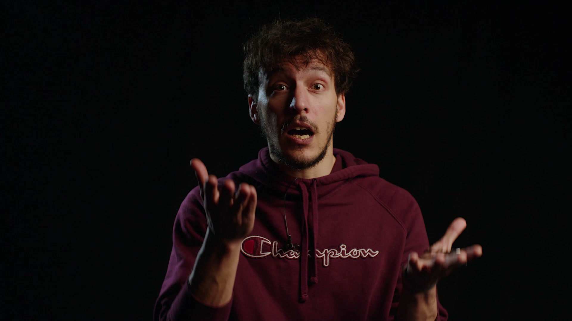 Interview participant in a burgundy hoodie gesturing against a dark background.